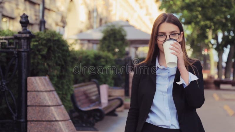 La mujer de negocios joven está caminando en la calle y bebe el café y smartphone con en la hora de la almuerzo
