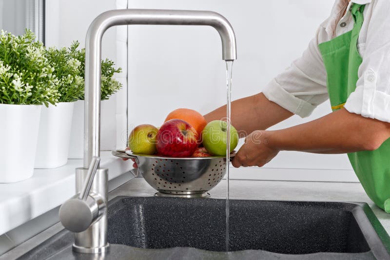 La mujer del cocinero lava las frutas debajo de la agua corriente de un golpecito de agua imagen de archivo La mujer del cocinero lava las frutas debajo de la agua corriente de un golpecito de agua imagen de archivo