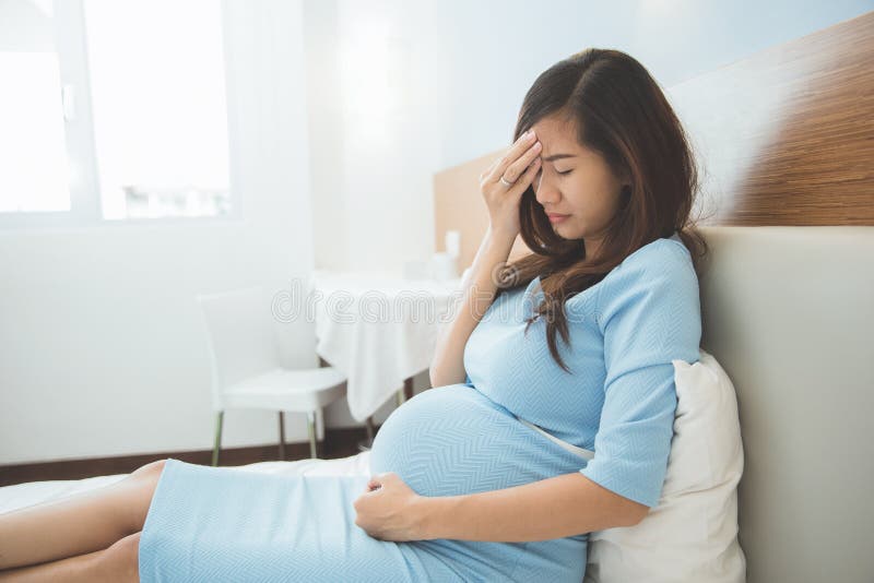 La mujer embarazada del asiático tiene dolor de cabeza que se sienta en su cama foto de archivo La mujer embarazada del asiático tiene dolor de cabeza que se sienta en su cama foto de archivo