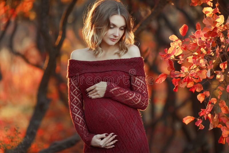 La mujer embarazada hermosa con el pelo rubio en vestido rojo largo y collar brillante se est? colocando en el bosque, sonriendo imagenes de archivo La mujer embarazada hermosa con el pelo rubio en vestido rojo largo y collar brillante se est? colocando en el bosque, sonriendo imagenes de archivo