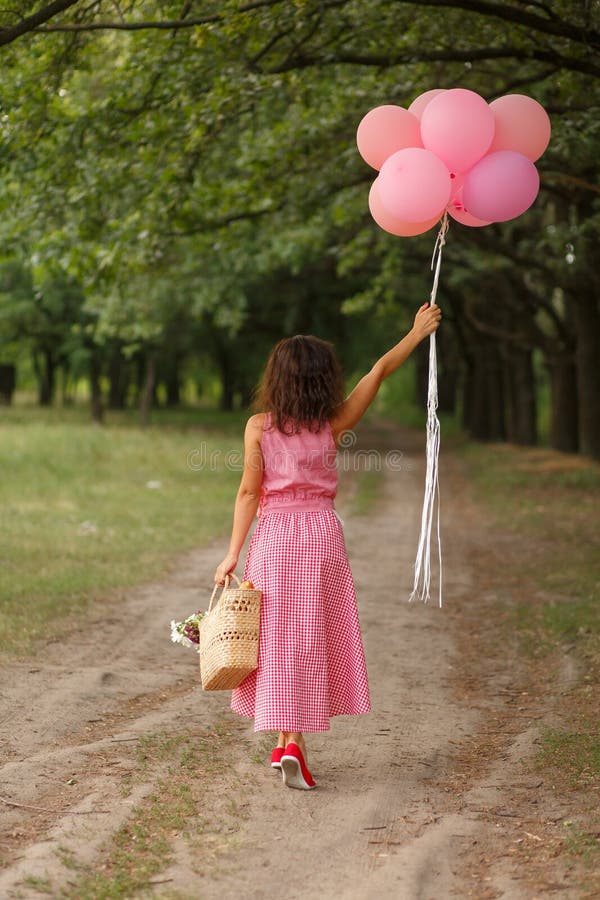 La mujer en vestido rosado con los globos rosados y la cesta de mimbre en domingo verde parquean imagen de archivo La mujer en vestido rosado con los globos rosados y la cesta de mimbre en domingo verde parquean imagen de archivo