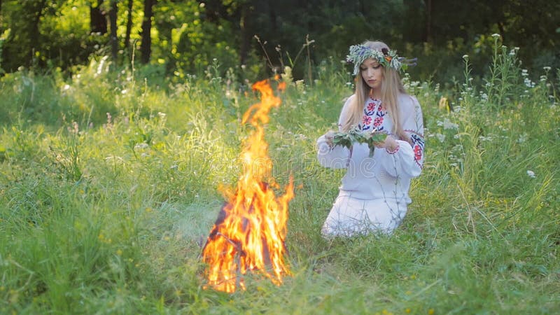 La mujer eslava en modelos bordados de una camisa, teje una guirnalda de las flores salvajes que se sientan por el fuego