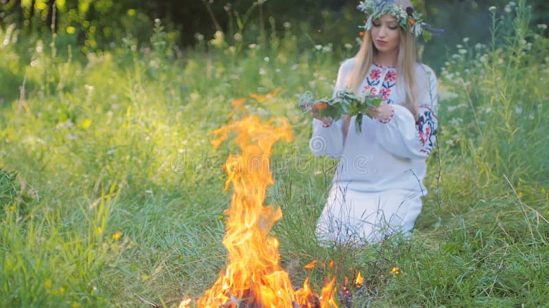 La mujer eslava en modelos bordados de una camisa, teje una guirnalda de las flores salvajes que se sientan por el fuego
