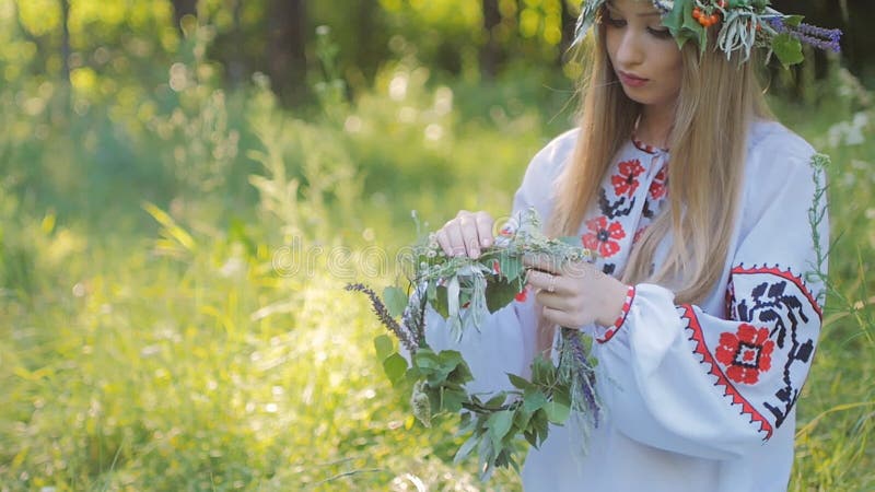 La mujer eslava en modelos bordados de una camisa, teje una guirnalda de las flores salvajes que se sientan por el fuego