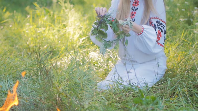 La mujer eslava en modelos bordados de una camisa, teje una guirnalda de las flores salvajes que se sientan por el fuego