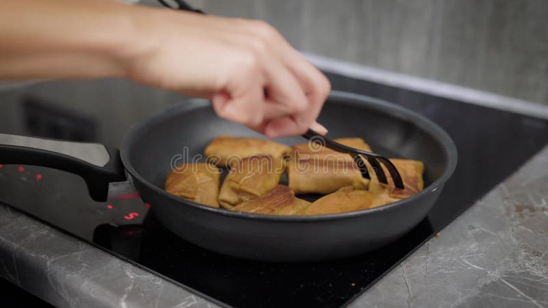 La mujer está asando las crepes con los rellenos en una cacerola en su electro cocina nacional, primer de manos