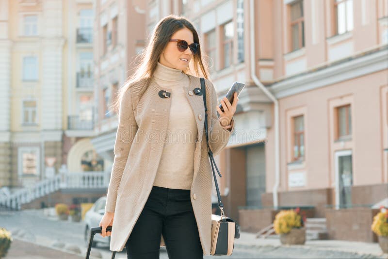 La mujer hermosa joven camina a lo largo de la calle de la ciudad con la maleta del viaje y el teléfono celular Sonrisa morena de imagen de archivo libre de regalías La mujer hermosa joven camina a lo largo de la calle de la ciudad con la maleta del viaje y el teléfono celular Sonrisa morena de imagen de archivo libre de regalías