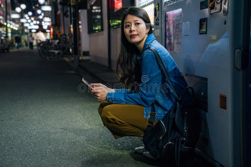 La mujer japonesa se agacha abajo por el amigo que espera de la máquina expendedora para señora elegante usando el app del contac imágenes de archivo libres de regalías La mujer japonesa se agacha abajo por el amigo que espera de la máquina expendedora para señora elegante usando el app del contac imágenes de archivo libres de regalías