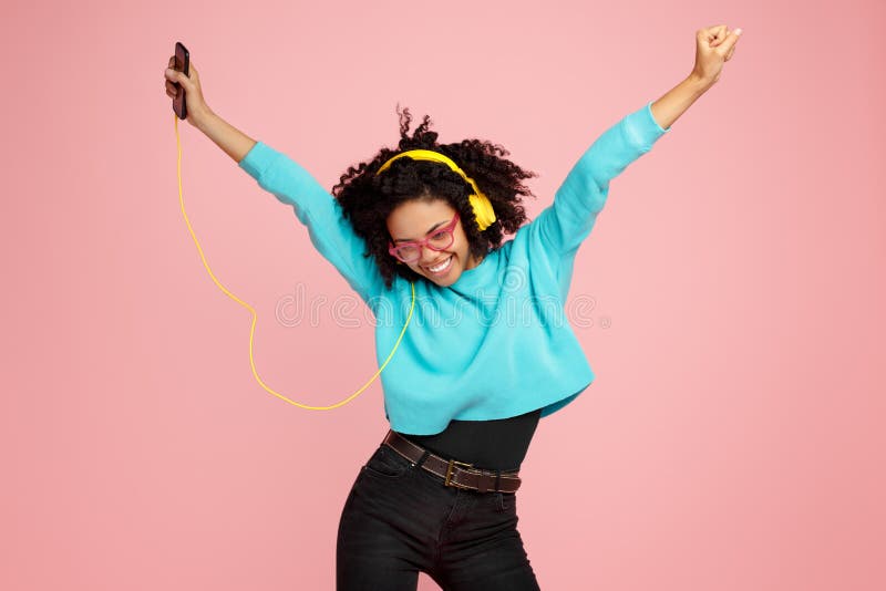 La mujer joven afroamericana bonita con sonrisa brillante vestida en ropa casual, los vidrios y los auriculares bailan sobre rosa fotos de archivo La mujer joven afroamericana bonita con sonrisa brillante vestida en ropa casual, los vidrios y los auriculares bailan sobre rosa fotos de archivo