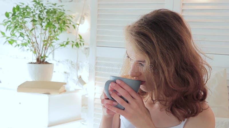 La mujer joven atractiva bebe cuidadosamente el café de una taza por la mañana