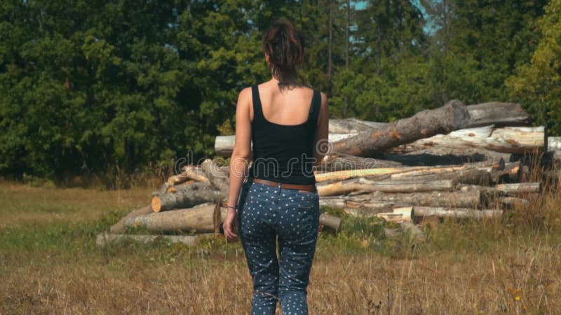 La mujer joven camina lentamente el campo de oro hacia registros y bosque conífero