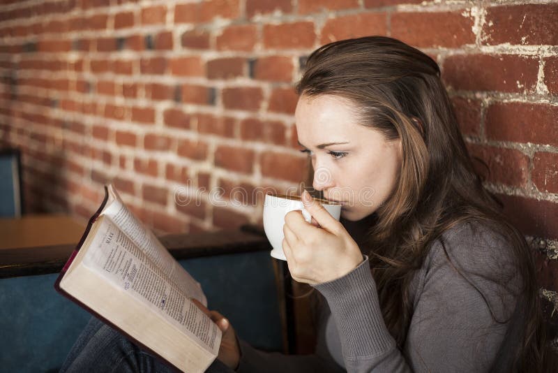 La mujer joven con la taza del café con leche lee su biblia
