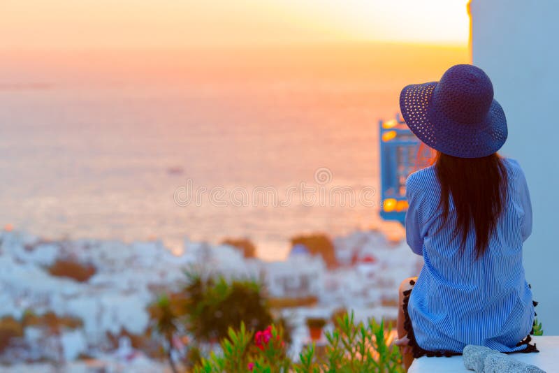 La mujer joven está mirando la puesta del sol sobre un mar en el pueblo famoso Mykonos con los edificios viejos en el fondo suave imagen de archivo libre de regalías La mujer joven está mirando la puesta del sol sobre un mar en el pueblo famoso Mykonos con los edificios viejos en el fondo suave imagen de archivo libre de regalías