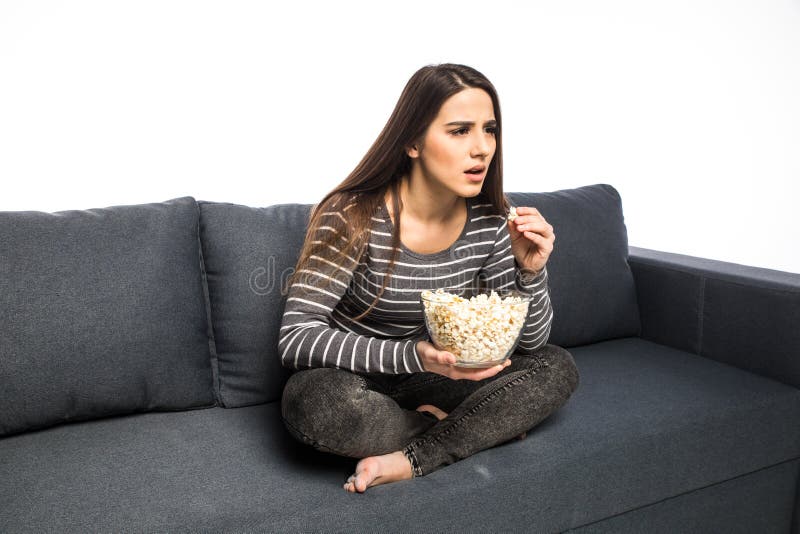 La mujer joven pasa su tiempo libre que ve la TV en el sofá que masca microprocesadores y el fondo del blanco de las palomitas imagen de archivo La mujer joven pasa su tiempo libre que ve la TV en el sofá que masca microprocesadores y el fondo del blanco de las palomitas imagen de archivo