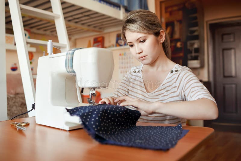 La mujer joven que cose con cose la máquina en casa mientras que se sienta por su lugar de trabajo El crear del diseñador de moda imagen de archivo La mujer joven que cose con cose la máquina en casa mientras que se sienta por su lugar de trabajo El crear del diseñador de moda imagen de archivo
