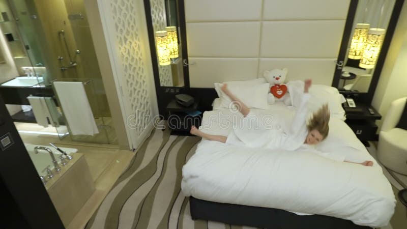 La mujer joven rubia hermosa feliz en una capa blanca sale del cuarto de baño, salta en la cama grande con el oso de peluche en c almacen de metraje de vídeo