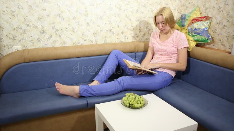 La mujer joven rubia se sienta en un sofá, lee el libro y come las uvas sabrosas almacen de metraje de vídeo