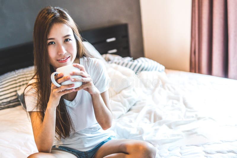 La mujer joven se sienta en la cama que bebe el café caliente fotos de archivo La mujer joven se sienta en la cama que bebe el café caliente fotos de archivo