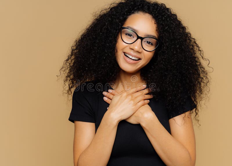 La mujer preciosa tocada con el peinado rizado, guarda las palmas en pecho, inclinaciones dirige, tiene sonrisa encantadora en ca foto de archivo libre de regalías La mujer preciosa tocada con el peinado rizado, guarda las palmas en pecho, inclinaciones dirige, tiene sonrisa encantadora en ca foto de archivo libre de regalías