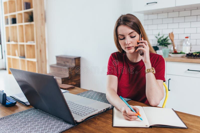 La mujer que trabaja en casa escribe notas mientras que habla en el teléfono fotografía de archivo