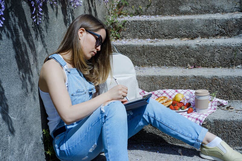 La mujer rubia joven del inconformista en gafas de sol dibuja en una tableta en su fotografía de archivo La mujer rubia joven del inconformista en gafas de sol dibuja en una tableta en su fotografía de archivo