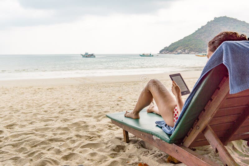 La mujer se sienta en sunbed en una playa tropical y lee un E-lector fotos de archivo libres de regalías La mujer se sienta en sunbed en una playa tropical y lee un E-lector fotos de archivo libres de regalías