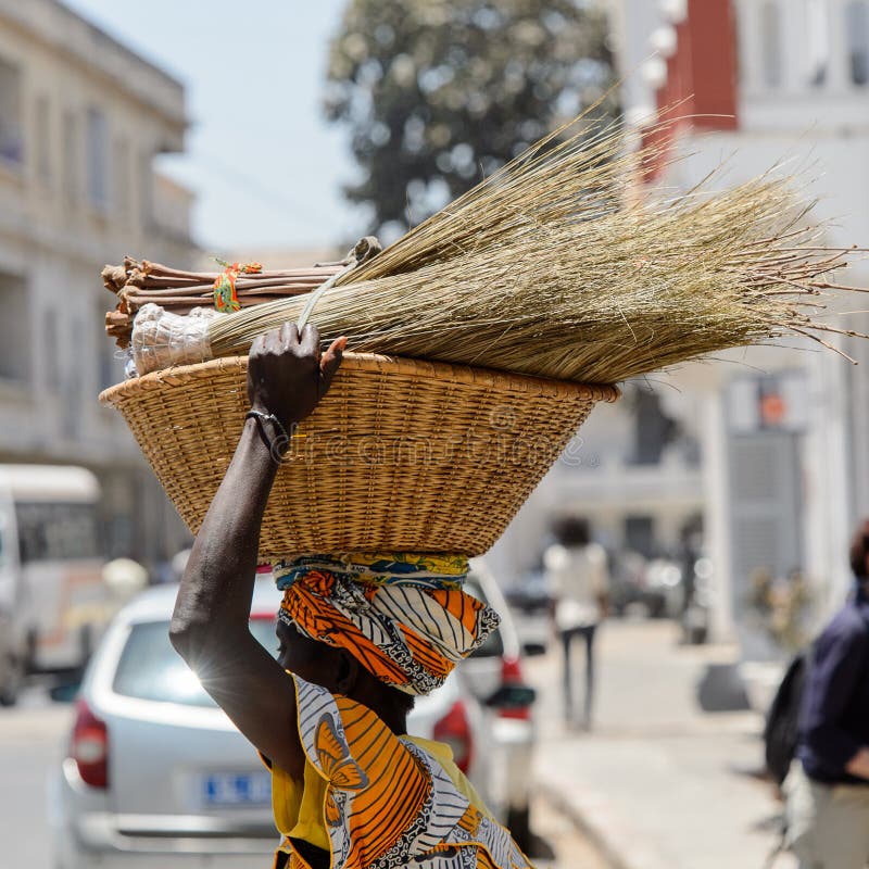 La mujer senegalesa no identificada lleva una cesta con las escobas en t imagen de archivo libre de regalías