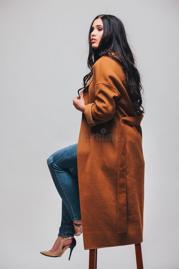La mujer sensual magnífica que se sienta en silla con el pelo recto oscuro lleva la capa elegante del brauwn fotos de archivo La mujer sensual magnífica que se sienta en silla con el pelo recto oscuro lleva la capa elegante del brauwn fotos de archivo