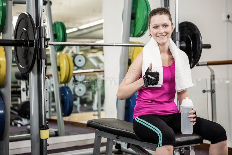 La mujer sonriente que se sienta en barbell bench y que muestra el pulgar para arriba imagen de archivo libre de regalías La mujer sonriente que se sienta en barbell bench y que muestra el pulgar para arriba imagen de archivo libre de regalías