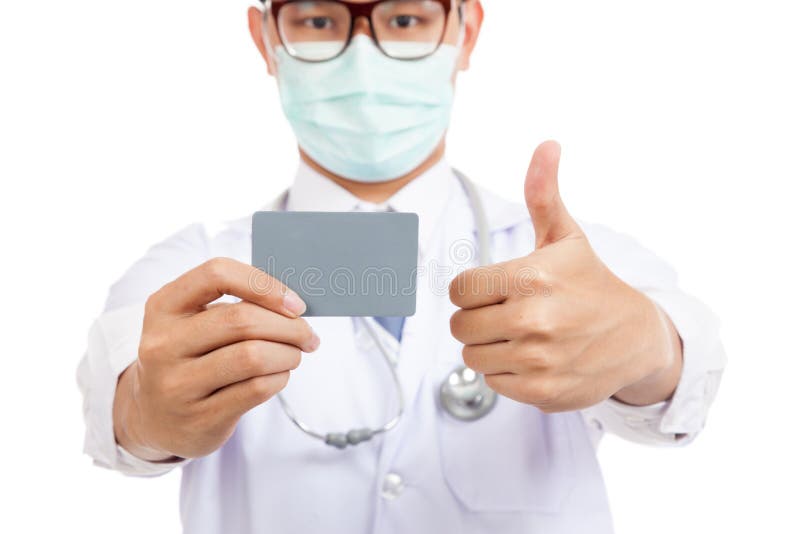 La máscara masculina asiática del desgaste del doctor manosea con los dedos para arriba con la tarjeta en blanco foto de archivo libre de regalías La máscara masculina asiática del desgaste del doctor manosea con los dedos para arriba con la tarjeta en blanco foto de archivo libre de regalías