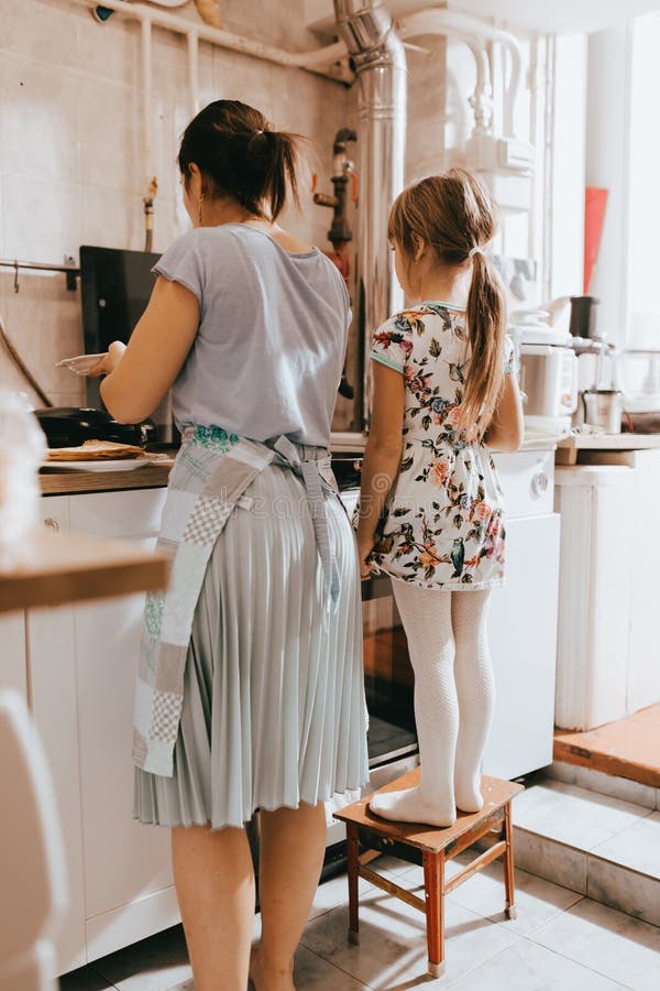 La niña que permanece en el taburete al lado de su madre está cocinando las crepes para el desayuno en la pequeña cocina acogedor imagen de archivo libre de regalías La niña que permanece en el taburete al lado de su madre está cocinando las crepes para el desayuno en la pequeña cocina acogedor imagen de archivo libre de regalías