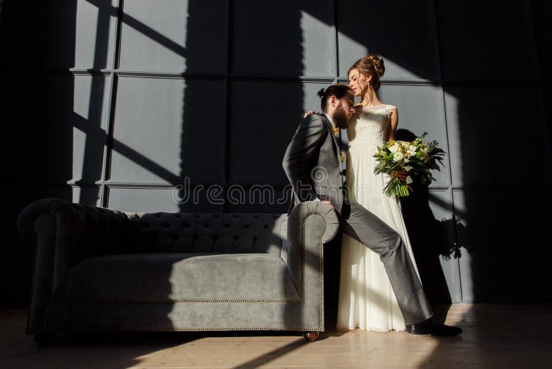 La novia está abrazando al novio que se sienta en el brazo del sofá Son encendidos por la luz dura de la ventana imagen de archivo La novia está abrazando al novio que se sienta en el brazo del sofá Son encendidos por la luz dura de la ventana imagen de archivo