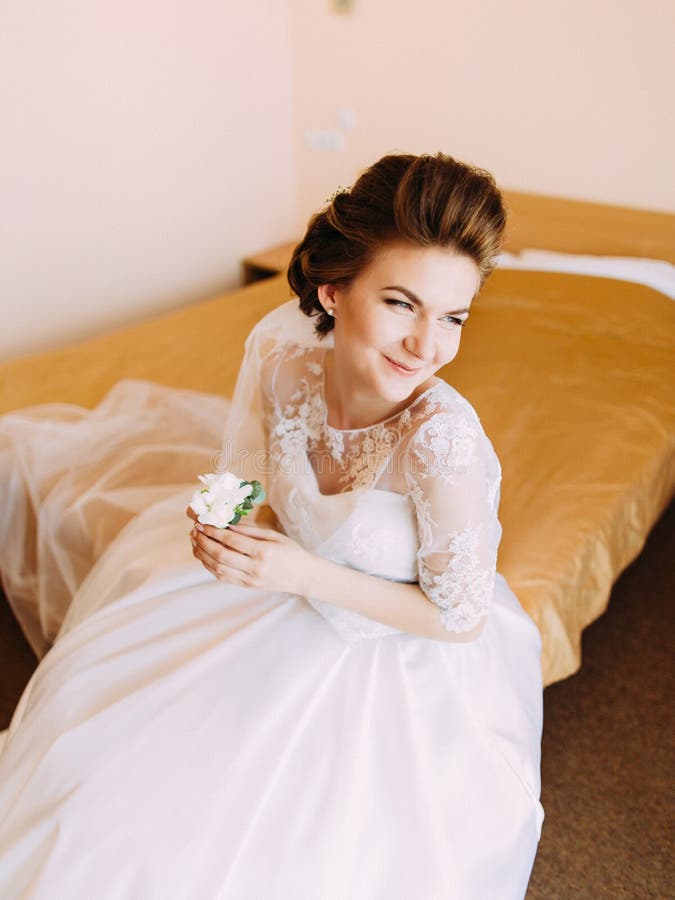 La novia que mira al lado derecho se está sentando en la cama y está sosteniendo el mini-ramo blanco fotografía de archivo La novia que mira al lado derecho se está sentando en la cama y está sosteniendo el mini-ramo blanco fotografía de archivo
