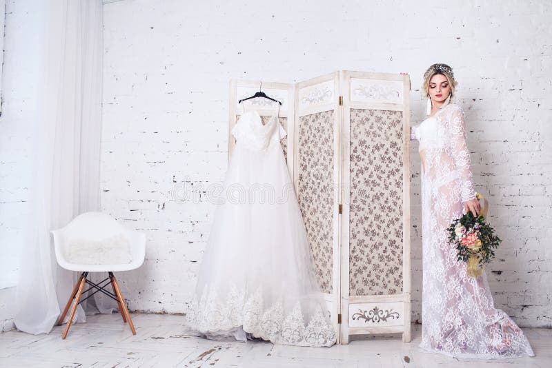 La novia, vestida en peignoir y ropa interior, coloca el escudo cercano con el vestido de boda y sostiene el ramo en sus manos foto de archivo libre de regalías La novia, vestida en peignoir y ropa interior, coloca el escudo cercano con el vestido de boda y sostiene el ramo en sus manos foto de archivo libre de regalías