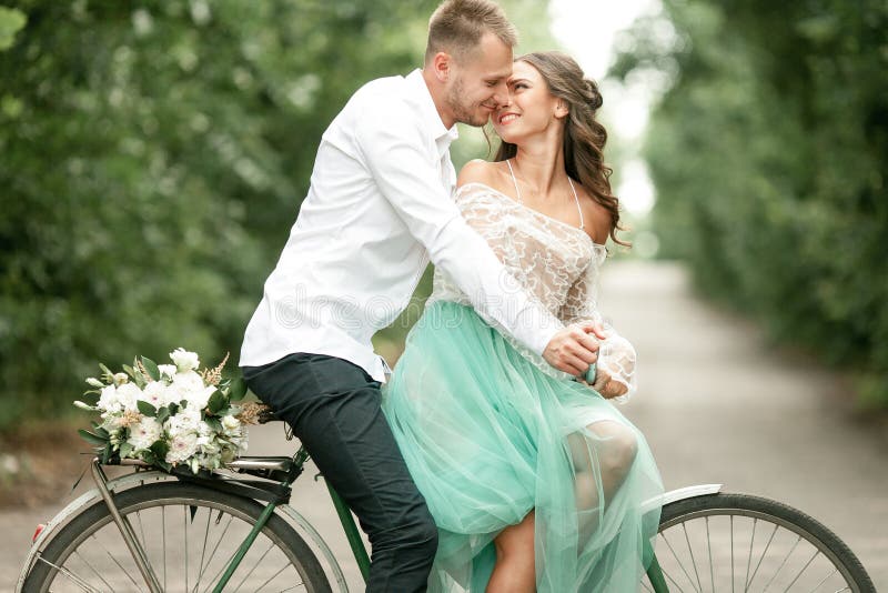 La novia y el novio se sientan en la bicicleta en el camino forestal, abrazan y sonríen imagen de archivo La novia y el novio se sientan en la bicicleta en el camino forestal, abrazan y sonríen imagen de archivo