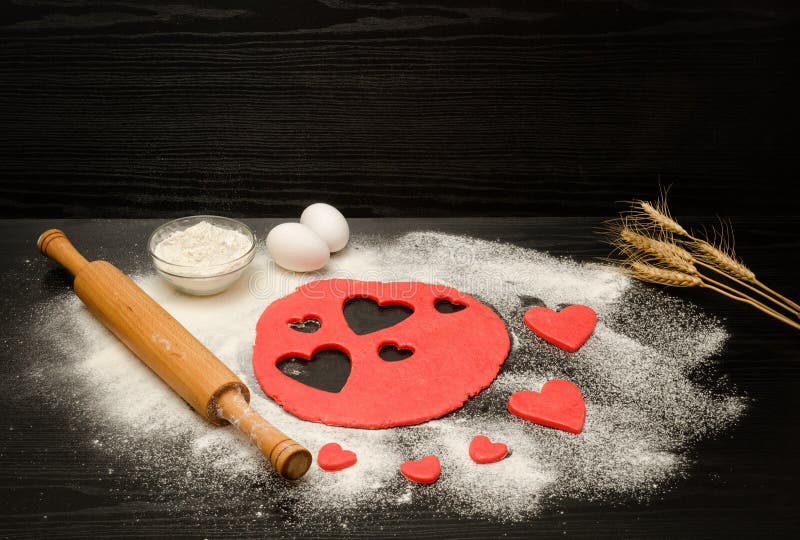 La pasta rossa, ha tagliato i cuori, la farina, le uova ed il matterello su un fondo nero, orecchie di grano, spazio per testo fotografia stock La pasta rossa, ha tagliato i cuori, la farina, le uova ed il matterello su un fondo nero, orecchie di grano, spazio per testo fotografia stock