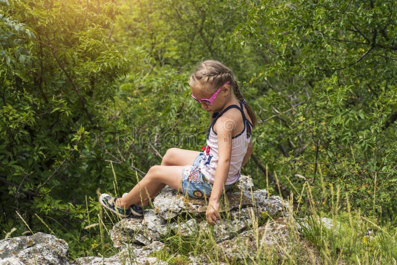 La pequeña muchacha brillante se sienta al borde de la montaña y mira en la distancia en las montañas imagen de archivo La pequeña muchacha brillante se sienta al borde de la montaña y mira en la distancia en las montañas imagen de archivo