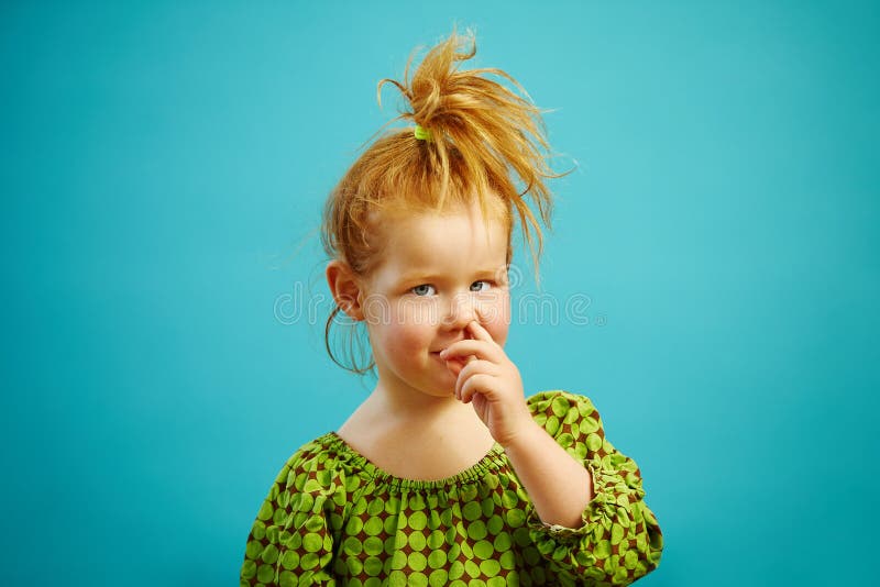 La pequeña muchacha linda del pelirrojo escoge su nariz con el finger en el azul aislado imagen de archivo libre de regalías La pequeña muchacha linda del pelirrojo escoge su nariz con el finger en el azul aislado imagen de archivo libre de regalías