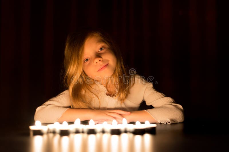 La petite fille blonde mignonne s'assied autour d'un bon nombre de bougie brûlante, au-dessus de fond foncé photos libres de droits La petite fille blonde mignonne s'assied autour d'un bon nombre de bougie brûlante, au-dessus de fond foncé photos libres de droits