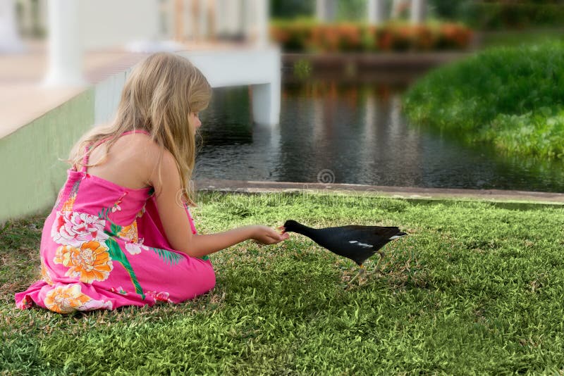 La petite fille caucasienne blonde alimente un oiseau dans un jardin tropical image libre de droits La petite fille caucasienne blonde alimente un oiseau dans un jardin tropical image libre de droits