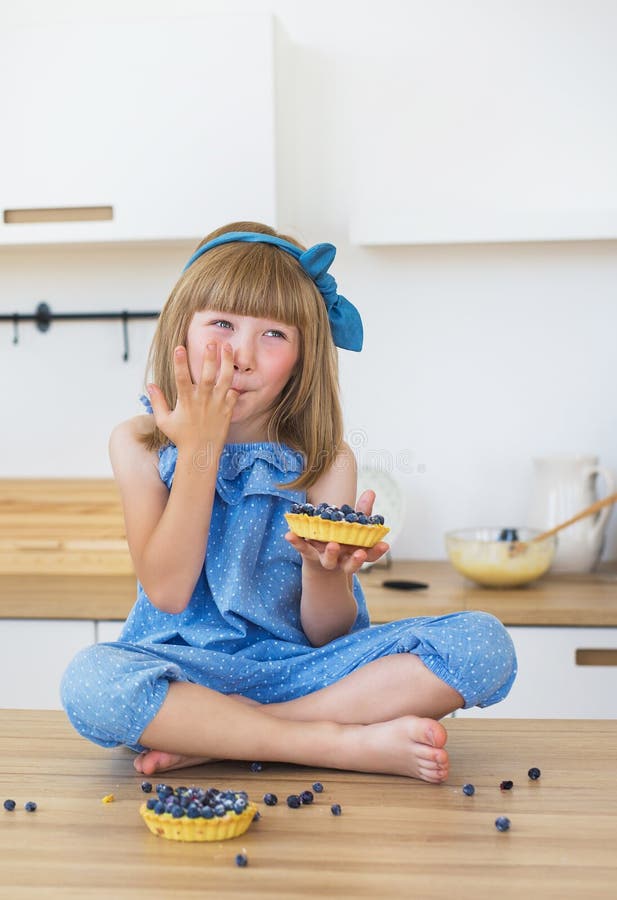 La petite fille mignonne dans la robe bleue mange un gâteau et lèche ses doigts photos libres de droits La petite fille mignonne dans la robe bleue mange un gâteau et lèche ses doigts photos libres de droits