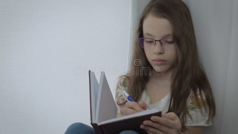 La petite fille sérieuse avec des verres écrit en son journal intime à la fenêtre