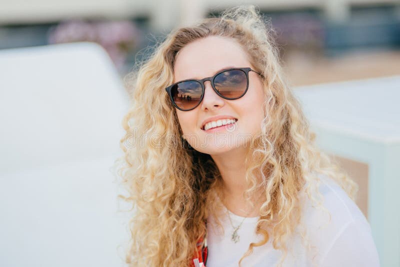 La photo de la jeune femelle attirante a les cheveux bouclés légers, porte les nuances à la mode, a le sourire toothy, montre les photos stock La photo de la jeune femelle attirante a les cheveux bouclés légers, porte les nuances à la mode, a le sourire toothy, montre les photos stock