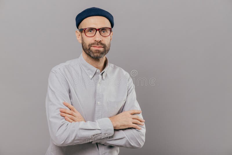 La photo du mâle sérieux maintient le coffre replié par bras, habillé dans la chemise formelle blanche, chapeau à la mode, pose p image stock La photo du mâle sérieux maintient le coffre replié par bras, habillé dans la chemise formelle blanche, chapeau à la mode, pose p image stock