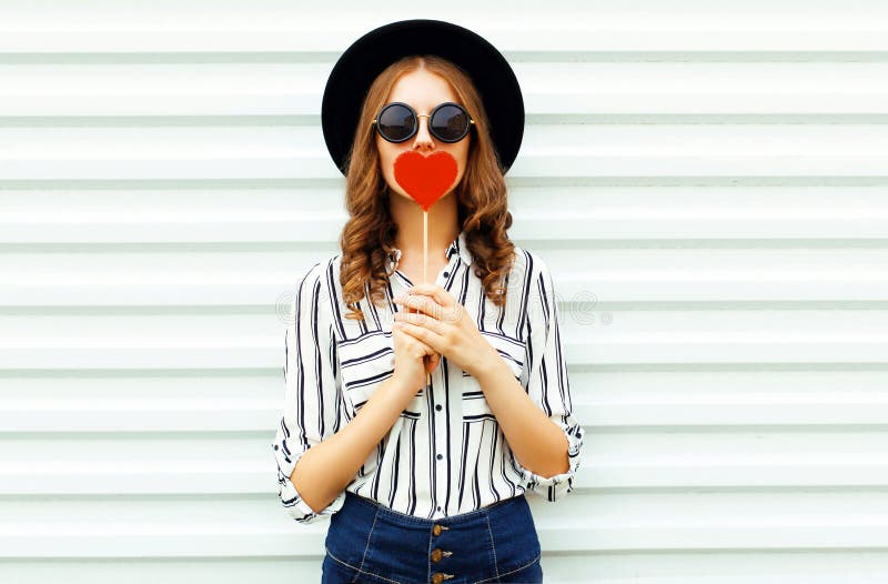 La piruleta en forma de corazón roja que se besa de la mujer joven del retrato u oculta sus labios en el sombrero redondo negro, fotografía de archivo libre de regalías La piruleta en forma de corazón roja que se besa de la mujer joven del retrato u oculta sus labios en el sombrero redondo negro, fotografía de archivo libre de regalías