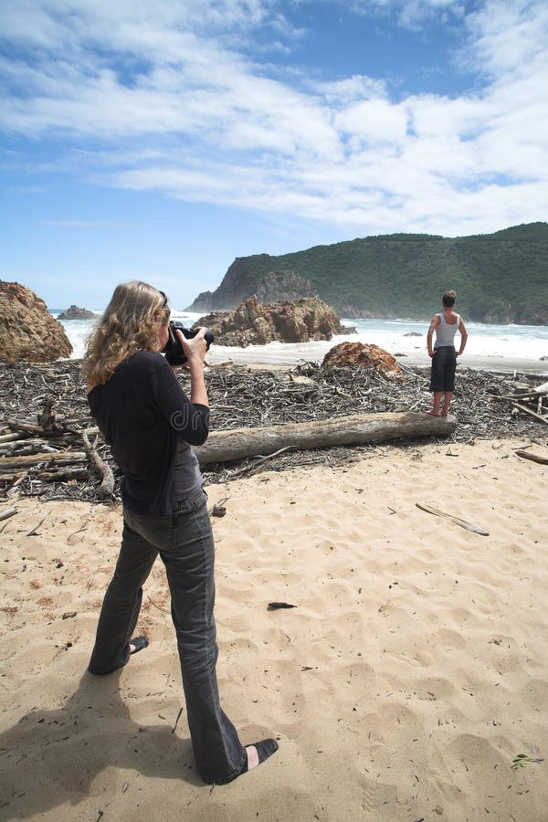 A model being shot on the beach at The Heads in Knysna, Western Cape, South Africa. The Female model is standing on some driftwood and flotsam with crashing waves in the background. The photographer is Female, wearing black. A model being shot on the beach at The Heads in Knysna, Western Cape, South Africa. The Female model is standing on some driftwood and flotsam with crashing waves in the background. The photographer is Female, wearing black.