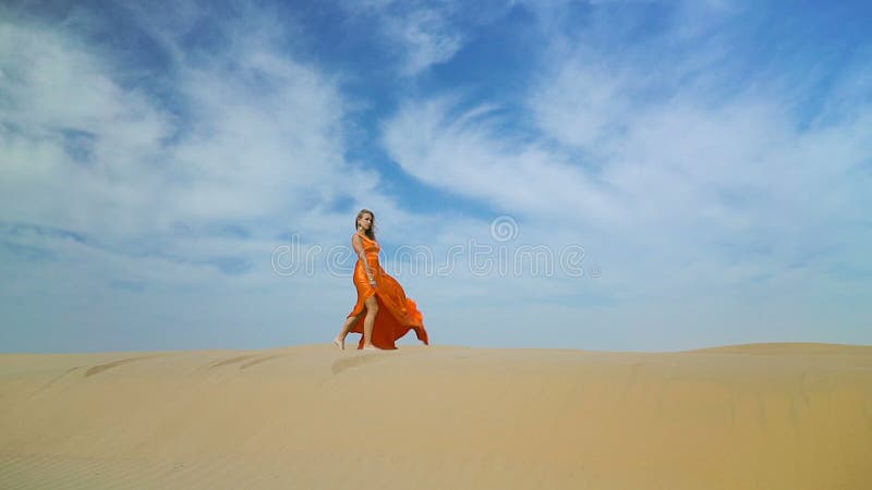 La ragazza cammina in deserto su barkhan La donna in vestito che cammina sulla sabbia-duna in deserto caldo sotto il sole, vento