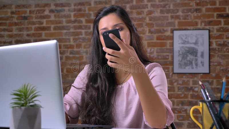 La ragazza casuale caucasica sveglia sta lavorando al computer portatile ed al DIS distratti con la telefonata, fronte sicuro ril