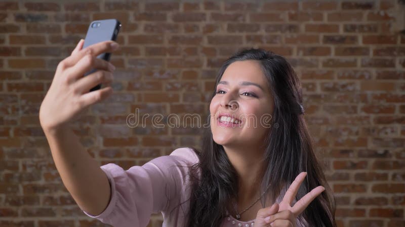 La ragazza caucasica mora felice di refrigerazione è selfies talike con il suo telefono, mostrante due dita e l'attimo sorridente