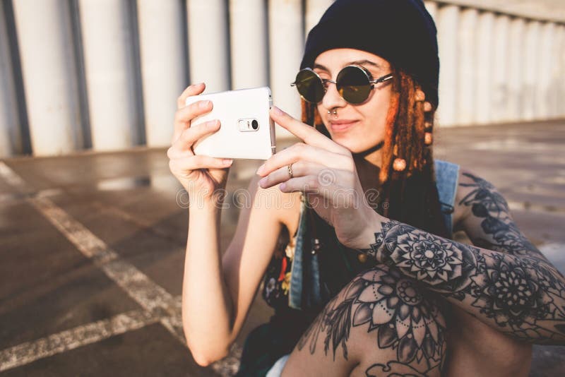 La ragazza con i tatuaggi ed i dreadlocks in un cappuccio blu utilizza uno smartphone contro lo sfondo di un muro di cemento immagine stock libera da diritti La ragazza con i tatuaggi ed i dreadlocks in un cappuccio blu utilizza uno smartphone contro lo sfondo di un muro di cemento immagine stock libera da diritti